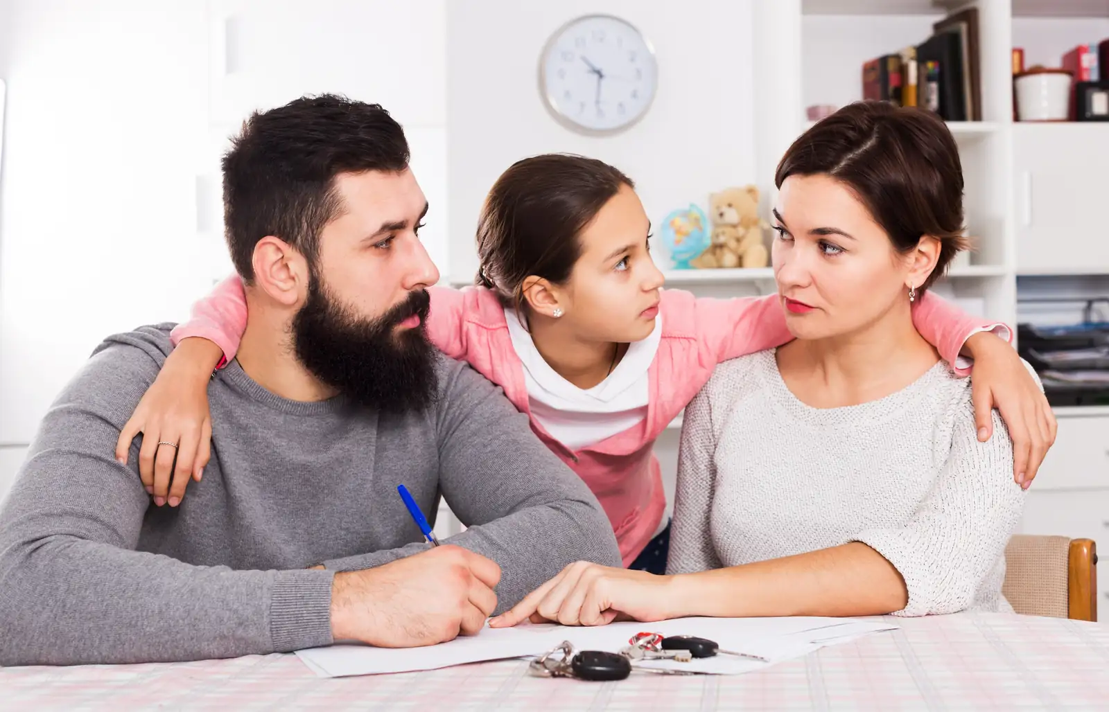 Parents seated at a table with paperwork while their daughter rests her arms around them