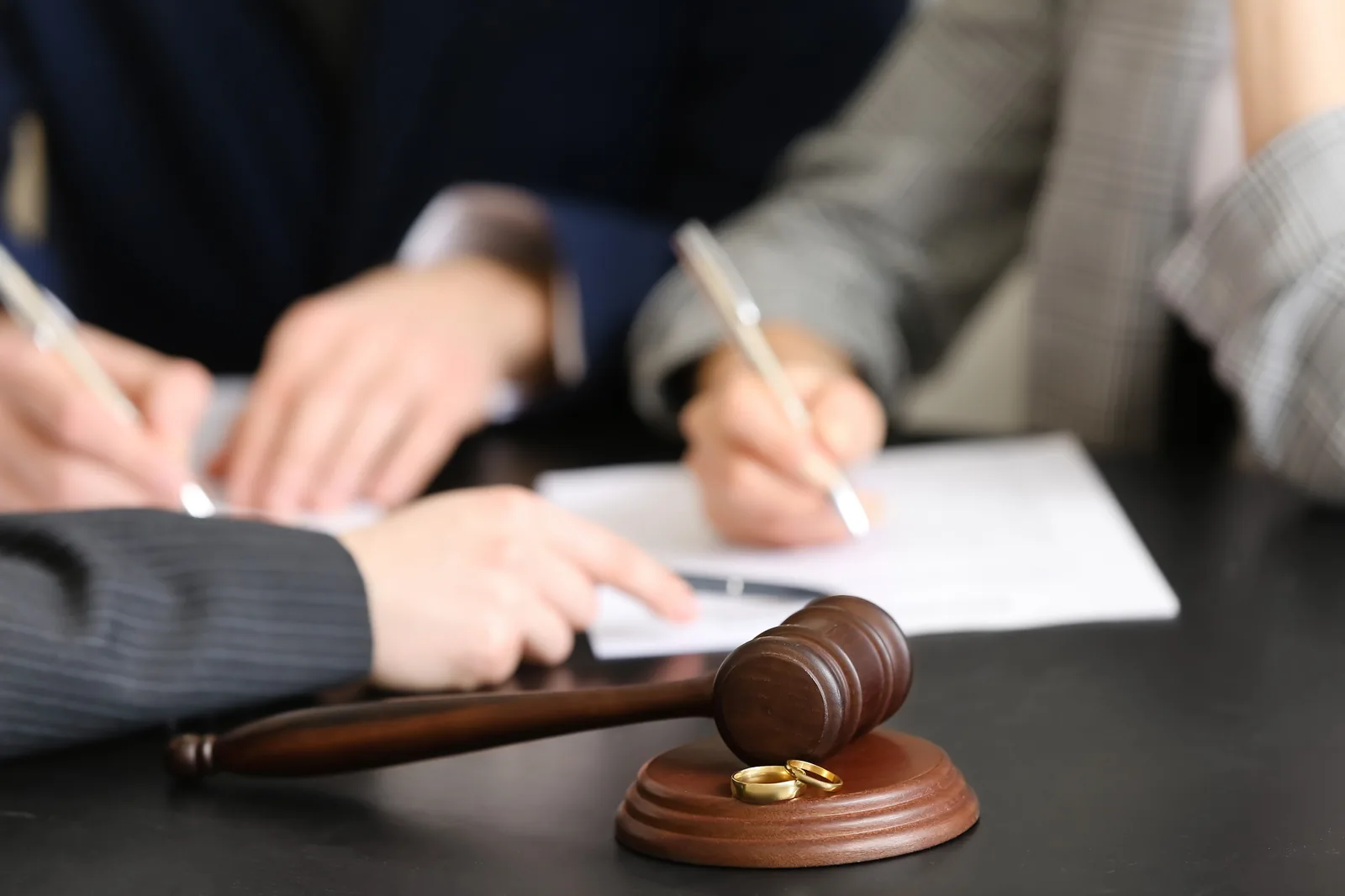Two people signing divorce paperwork at a table beside a judge's gavel and wedding rings